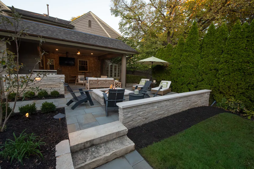 Paver terrace with steps, low stone wall, covered patio, lawn edge, and planting beds